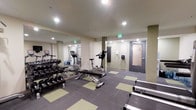 This single-level fitness room offers a practical and functional workout space designed to accommodate a range of exercise needs. The room features a mirrored wall along one side, which visually expands the space and allows users to monitor their form during workouts. Recessed ceiling lighting provides consistent, even illumination throughout the area.

The free weights section includes a multi-tiered dumbbell rack stocked with a variety of weights, accompanied by a flat adjustable bench, making it suitable for strength training exercises. Cardio equipment includes at least one upright stationary bike and an additional cardio machine visible in the mirrored reflection, both from the True fitness brand. The flooring consists of a combination of gray and light green interlocking tiles, offering cushioning and traction during workouts.

A wall-mounted equipment holder and informational signage are positioned near the entrance, adding organizational functionality. The room is accessed through a sturdy gray door equipped with a glass panel. An emergency exit sign is clearly visible, ensuring safety compliance. Overall, the space is clean, organized, and purpose-built for efficient fitness use.