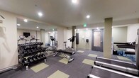 This single-level fitness room offers a practical and functional workout space designed to accommodate a range of exercise needs. The room features a mirrored wall along one side, which visually expands the space and allows users to monitor their form during workouts. Recessed ceiling lighting provides consistent, even illumination throughout the area.

The free weights section includes a multi-tiered dumbbell rack stocked with a variety of weights, accompanied by a flat adjustable bench, making it suitable for strength training exercises. Cardio equipment includes at least one upright stationary bike and an additional cardio machine visible in the mirrored reflection, both from the True fitness brand. The flooring consists of a combination of gray and light green interlocking tiles, offering cushioning and traction during workouts.

A wall-mounted equipment holder and informational signage are positioned near the entrance, adding organizational functionality. The room is accessed through a sturdy gray door equipped with a glass panel. An emergency exit sign is clearly visible, ensuring safety compliance. Overall, the space is clean, organized, and purpose-built for efficient fitness use.