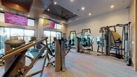 This single-level fitness center is designed with both functionality and aesthetics in mind, offering a well-rounded exercise environment suited for a variety of workout routines. The space features warm wood-paneled accent walls that lend a contemporary yet inviting atmosphere, complemented by patterned carpet flooring with orange and gray tones that adds visual interest underfoot.

The cardio zone is equipped with a row of TRUE-brand treadmills and multiple elliptical and cycling machines, all positioned to face large windows that bring in natural light and outdoor views. A wall-mounted flat-screen television provides entertainment during workouts, while a ceiling fan adds air circulation to the space.

The strength training area includes Hoist cable and multi-use machines, a set of dumbbells neatly arranged on a tiered rack near the entrance, and a dedicated functional training zone stocked with stability balls, medicine balls, resistance bands, foam rollers, and an adjustable weight bench. Several large round and rectangular mirrors are strategically placed throughout to allow users to monitor their form.

The entrance features double glass doors framed by wood paneling, and a built-in counter area with shelving provides a convenient station near the entry. Recessed lighting throughout ensures the space is well-illuminated at all times.
