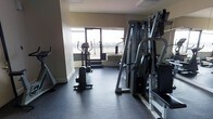 This fitness center is a dedicated exercise space designed to support a well-rounded workout routine. The room features durable rubber flooring throughout, providing a safe and functional surface suited for both cardio and strength training activities. A large window dominates one wall, flooding the space with natural light and offering views of the surrounding outdoor landscape, including a balcony or terrace area visible beyond the glass.

The equipment selection is comprehensive and thoughtfully arranged. Cardio machines, including elliptical trainers and what appears to be a stair climber or treadmill, are positioned near the window to take advantage of the natural light and views. On the opposite side of the room, a robust multi-station cable and weight machine provides a wide range of strength training options, accommodating various exercise routines for users of different fitness levels.

The neutral-toned walls and practical layout contribute to a clean, focused atmosphere. The space is efficiently organized to allow freedom of movement between equipment stations. Overall, this fitness center offers a functional and well-lit environment that combines essential exercise equipment with a pleasant, light-filled setting conducive to an active and healthy lifestyle.