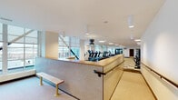 Located at 3850 Boston St in Baltimore, MD, this fitness center offers a thoughtfully designed, single-floor workout facility that balances functionality with a clean, modern aesthetic. The space is organized to accommodate a wide range of fitness activities, with distinct zones dedicated to cardio, strength training, and free weights.

The cardio area features a row of treadmills equipped with personal screens, alongside elliptical trainers and a rowing machine, all positioned near large windows that allow natural light to fill the space. The strength training zone includes a multi-station cable and weight machine, as well as a power rack, providing options for a full-body workout. A dedicated free weights section houses an extensive dumbbell rack flanked by floor-to-ceiling mirrors that enhance the sense of openness.

Throughout the facility, the flooring consists of a distinctive multi-tone checkered tile pattern in muted greens, tans, and grays, contributing to the space's contemporary character. Circular flush-mount and cylindrical pendant light fixtures provide consistent, even illumination across all areas. Tiled half-walls with wood-capped partitions add architectural interest near the entry, accompanied by a simple bench seating area. The overall ambiance is bright, open, and professionally appointed, making it a well-rounded fitness destination.