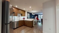 This contemporary single-floor residence showcases a thoughtfully designed open-concept layout that seamlessly connects the kitchen, dining, and living areas. The kitchen is well-appointed with warm walnut-toned cabinetry, white quartz countertops, a full suite of stainless steel appliances including a gas range, built-in microwave, dishwasher, and refrigerator, complemented by under-cabinet lighting and a clean white tile backsplash. Pendant lights above the peninsula add a modern touch, while wood-look flooring flows throughout the main living area. The living space makes a bold design statement with a deep navy accent wall, a white sofa, and a striking red egg-style chair, lending the room a vibrant, modern character. French doors at the far end of the living area invite natural light and provide an outdoor connection. The bedroom features a dramatic dark navy accent wall, carpeted flooring, and a window overlooking greenery, creating a calm and restful retreat. A second room serves as a flexible sleeping or study space with bright natural light. The bathroom is finished with large-format grey tile, a tub and shower combination with sliding glass doors, a wood-toned vanity, and crisp white surfaces, creating a clean and functional space.