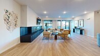 Located at 705 1st Ave SW in Rochester, Minnesota, this clubroom and sports simulator facility offers a well-appointed, single-floor amenity space designed for both relaxation and active recreation. The main lounge area is generously sized, featuring warm light wood flooring, large black-framed windows that invite abundant natural light, and a striking dark-tiled fireplace flanked by a wall-mounted television. Comfortable seating arrangements with contemporary furnishings create an inviting social atmosphere throughout the open layout. Adjacent to the lounge is a sleek kitchenette outfitted with white cabinetry, white quartz countertops, a stainless steel sink with a gold-tone faucet, and a bold navy blue vertical tile backsplash that adds a vibrant design focal point. Open metal shelving and a built-in microwave complete the functional space. The sports simulator room is a standout feature, housing a Full Swing simulator with a large projection screen, dark carpet turf flooring, and ample space for equipment storage. A dedicated viewing and lounge area adjacent to the simulator includes patterned carpet, a long communal table, and curated wall art with a golf theme, providing a comfortable space to watch and socialize.