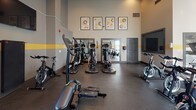 This fitness center is designed to support a comprehensive workout experience within a single open floor. The space features high ceilings that enhance the sense of openness, with a distinctive yellow accent stripe running along the perimeter walls, complemented by a neutral gray color palette and durable rubber flooring throughout. Recessed lighting and track lighting illuminate the interior, while large windows along the exterior walls allow ample natural light to flow in, offering views of the outdoors. The cardio equipment selection is extensive, including multiple treadmills, spin bikes, rowing machines, and elliptical trainers, providing users with a wide variety of workout options. A dedicated wall of mirrors expands the visual space and allows users to monitor their form during exercise. Wall-mounted flat-screen TVs are positioned for easy viewing from multiple workout stations. Decorative framed artwork adorns the upper walls, adding a polished, finished aesthetic. The space also includes a yoga or exercise mat for floor-based workouts, and a sanitation station with a waste receptacle supports cleanliness and hygiene. Multiple entry and exit points, including a glass door opening to an outdoor area, contribute to easy accessibility and natural ventilation.
