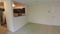 This single-floor residence offers a clean, functional layout designed for comfortable everyday living. The living area is finished with wall-to-wall neutral carpeting that adds warmth underfoot, while crisp white walls provide a bright and versatile backdrop throughout the space. A pass-through opening connects the living area to the kitchen, creating an open, airy feel that encourages interaction between the two spaces. The kitchen is well-appointed with rich, dark espresso-toned cabinetry that offers generous storage, paired with stainless steel appliances including a microwave and refrigerator. A dark stone or granite-style countertop runs along the kitchen's pass-through bar, providing additional prep and casual dining space. Recessed and flush-mount ceiling lighting ensures the interiors remain well-lit. The overall ambiance is clean and understated, with a neutral color palette that makes the space feel open and move-in ready. The straightforward, single-level layout makes the unit easily navigable, and the seamless flow between the kitchen and living areas makes it well-suited for both daily living and entertaining.