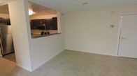 This single-floor residence offers a clean, functional layout designed for comfortable everyday living. The living area is finished with wall-to-wall neutral carpeting that adds warmth underfoot, while crisp white walls provide a bright and versatile backdrop throughout the space. A pass-through opening connects the living area to the kitchen, creating an open, airy feel that encourages interaction between the two spaces. The kitchen is well-appointed with rich, dark espresso-toned cabinetry that offers generous storage, paired with stainless steel appliances including a microwave and refrigerator. A dark stone or granite-style countertop runs along the kitchen's pass-through bar, providing additional prep and casual dining space. Recessed and flush-mount ceiling lighting ensures the interiors remain well-lit. The overall ambiance is clean and understated, with a neutral color palette that makes the space feel open and move-in ready. The straightforward, single-level layout makes the unit easily navigable, and the seamless flow between the kitchen and living areas makes it well-suited for both daily living and entertaining.
