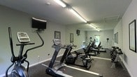 This fitness center is a dedicated, single-floor workout facility designed to support a well-rounded exercise routine. The space is outfitted with a lineup of cardio equipment, including treadmills and stationary bikes, arranged to allow comfortable movement between machines. A mirrored wall runs along one side of the room, visually expanding the space while allowing users to monitor their form during workouts. Rubber flooring covers the entire area, providing durability and impact absorption suited for active use. Overhead fluorescent lighting ensures the space is consistently and evenly illuminated throughout. Multiple wall-mounted flat-screen televisions are positioned at convenient viewing angles, adding an element of entertainment during workouts. Informational boards are mounted on the walls, likely displaying workout guides or facility guidelines. The walls are finished in a neutral tone, contributing to a clean and focused environment. A window near the rear of the space allows some natural light to enter, providing a connection to the outdoors. The overall layout is practical and efficiently organized, making the most of the available footprint to deliver a functional and accessible fitness environment.