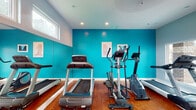 Located at Ashton Park in Louisville, Kentucky, this dedicated fitness center offers residents a clean and energizing space designed to support a variety of workout routines. The facility is single-level and thoughtfully laid out to maximize usability throughout.

The main exercise area is anchored by bold teal accent walls, full-length wall mirrors, and warm hardwood flooring that lend the space a vibrant yet functional character. Recessed lighting ensures the room is well-lit at all hours. The cardio section is outfitted with multiple treadmills and an elliptical machine, providing ample options for cardiovascular training without crowding.

A separate area houses a multi-function cable strength machine along with stability exercise balls, accommodating strength training and functional fitness exercises. A wall-mounted flat-screen television is positioned for easy viewing during workouts, adding an entertainment element to the experience.

Natural light filters into the space through horizontal windows positioned high along the walls, keeping the atmosphere bright and open. The entry area features soft blue-gray walls, a glass-panel door with exterior views, and posted fitness center guidelines, giving the space a professional and organized feel. The overall ambiance is clean, modern, and motivating — a practical amenity designed to serve an active community.