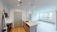 This single-floor unit presents a clean, contemporary design with a cohesive neutral palette and thoughtful layout. The open-concept kitchen and living area serves as the heart of the space, where light hardwood-style flooring runs underfoot and a large window allows natural light to fill the room. The kitchen is well-appointed with gray flat-panel cabinetry extending to the ceiling, stainless steel appliances including a refrigerator, electric range, and built-in microwave, and a white quartz countertop. A kitchen island with a stainless steel sink and pendant lighting above provides both additional prep space and a natural visual anchor between the kitchen and living area. Paneled gray interior doors are used consistently throughout, contributing to a unified aesthetic. The bedroom features neutral carpet flooring and a generously sized window overlooking the surrounding exterior. The bathroom includes a glass-enclosed shower, white tile surround, a vanity with quartz countertop, and gray cabinetry matching the kitchen. A separate utility or laundry area offers additional cabinet storage and counter space. Recessed lighting and clean white walls reinforce the modern, move-in-ready character of the space.