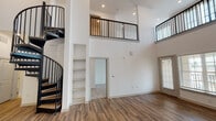 This two-story space is defined by its dramatic double-height living area, where soaring ceilings and recessed lighting create an open, airy atmosphere. A striking black spiral staircase serves as both a functional and architectural centerpiece, connecting the main level to an upper-floor mezzanine enclosed by sleek black metal railings. The main level features warm hardwood-style flooring throughout the open-plan living and kitchen areas. Built-in shelving is integrated into the living space, providing practical storage without compromising the clean aesthetic.

The kitchen is well-appointed with taupe cabinetry, granite countertops, stainless steel appliances, and a kitchen island with pendant lighting above, making it a functional and visually appealing workspace.

The upper level hosts carpeted bedrooms, each offering natural light through generously sized windows. One bedroom benefits from tall ceilings with elevated windows, adding a sense of height and brightness. Both levels include walk-in closets outfitted with wire shelving systems for organized storage. The bathroom features a granite-topped vanity with dark cabinetry, a large mirror, tiled flooring, and a tub-shower combination, reflecting a consistent modern design language found throughout the space.