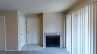 This single-story space presents a straightforward and functional layout centered around a welcoming living area. The room features a tile-surround fireplace with a black glass front, serving as a natural focal point along the main wall. The flooring transitions between tile near the entryway and carpet in the living area, offering a practical and comfortable combination underfoot.

Natural light enters the space through a large sliding glass door fitted with vertical blinds, brightening the interior and providing access to the outdoors. The entryway is defined by a solid white front door with multiple lock points, conveying a sense of security and practicality.

The walls are painted in a neutral beige tone, complementing the light-colored tile flooring and creating a clean, understated aesthetic throughout. The ceiling features a textured popcorn finish, consistent with the overall style of the space.

The layout is open and unfurnished, allowing for flexible arrangement of furniture and décor. Overall, the space offers a simple, functional environment with key comforts such as the fireplace and natural light, making it a versatile and livable setting.