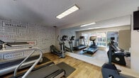 Located at 21701 Foothill Blvd in Hayward, California, this single-floor fitness center is a well-equipped exercise facility designed to support a well-rounded cardio workout routine. The room features durable hardwood-style flooring throughout, providing a clean and practical surface for exercise equipment and movement.

The space is outfitted with multiple treadmills, elliptical trainers, and a recumbent stationary bike, offering a variety of cardiovascular training options. A wall-mounted flat-screen television is positioned for convenient viewing during workouts. Large floor-to-ceiling mirrors line one wall, visually expanding the space and allowing users to monitor their form while exercising.

One of the room's most distinctive design elements is a bold, typography-style accent wall featuring fitness-related keywords such as "Wellness," "Athletic," "Body," and "Strength," lending the space an energetic and motivating atmosphere. Flush-panel recessed lighting and a textured ceiling complete the interior aesthetic.

Natural light flows generously through large windows and a glass door that opens directly to the outdoor pool area, where patio seating and lush greenery are visible. This connection to the outdoors enhances the overall ambiance of the fitness center, creating a bright and inviting environment conducive to an active lifestyle.