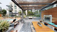 This expansive outdoor amenity space is thoughtfully designed to deliver a resort-like experience within a contemporary urban setting. At its centerpiece is a generously sized swimming pool with clear turquoise water, complemented by an adjacent spa. The pool deck is finished in large-format porcelain tile and lined with cushioned chaise lounges and patio umbrellas, providing ample space for sunbathing and relaxation.

A substantial pergola structure with warm wood slat detailing anchors a dedicated lounge and dining zone. Beneath it, a teak dining table with white chairs sits adjacent to an outdoor kitchen equipped with a gas grill, ideal for al fresco entertaining. A wall-mounted media screen and fireplace add functional character to this covered area.

The surrounding landscape features raised wood-clad planters filled with ornamental grasses, bamboo, and drought-tolerant plantings, creating natural privacy screens and a lush aesthetic. Additional seating nooks with teal accent chairs and daybed loungers offer quieter retreats throughout the courtyard. The entire space is framed by the building's modern façade, reinforcing its cohesive, contemporary design character.