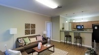 Located at 105 Doleac Dr. in Hattiesburg, Mississippi, this single-floor space offers a thoughtfully designed open-concept layout that seamlessly connects the living and kitchen areas. The living room is generously sized, featuring warm hardwood-style flooring, neutral-toned walls, and elegant crown molding that adds a refined architectural touch. A flush ceiling light fixture provides bright, even illumination, while the open flow of the space creates an inviting and airy atmosphere. Decorative wall art and ample room for furnishings make this area both functional and welcoming. The kitchen is positioned adjacent to the living area and is highlighted by a raised breakfast bar with granite-style countertops, ideal for casual dining or entertaining. Warm wood cabinetry lines the kitchen walls, complemented by stylish pendant lighting that hangs above the bar. The pass-through design of the kitchen enhances the open feel of the entire living space. Crown molding continues throughout, maintaining a consistent and polished aesthetic. The unit's single-floor layout ensures convenient and accessible navigation throughout, making it a practical and attractive space in the heart of Hattiesburg.