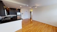 This single-floor space features an open-concept design that integrates the kitchen and living area into one cohesive, light-filled environment. Warm-toned hardwood floors extend throughout, lending a polished and inviting aesthetic to the entire space.

The kitchen is positioned to the left and is outfitted with dark espresso cabinetry that contrasts sharply against the dark granite countertops and stainless steel refrigerator. A peninsula counter provides a natural boundary between the kitchen and the living area, offering both additional prep space and a casual seating opportunity.

The living area is generously proportioned, benefiting from a clean, minimalist design with crisp white walls and a track lighting system mounted to the ceiling, offering directed and adjustable illumination. Recessed lighting in the kitchen adds further depth and brightness to the overall space.

A solid entry door anchors the far wall, flanked by a hallway that leads to the private areas of the unit, including what appears to be a bedroom. The straightforward, open layout ensures excellent sightlines throughout, making the space feel expansive and functional.
