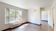 This single-floor unit offers a thoughtfully organized layout with consistent hardwood-style flooring and neutral white walls throughout, creating a clean and cohesive aesthetic. The galley kitchen is well-equipped with light wood cabinetry lining both sides, ample counter space, a double sink, a dishwasher, a full-size refrigerator, and an electric range with oven — all in white. Overhead fluorescent lighting keeps the kitchen bright and functional.

The living areas are open and airy, benefiting from large windows that invite abundant natural light and offer views of surrounding greenery. A wall-mounted HVAC unit provides climate control, and baseboard heating is present in multiple rooms. The open flow between spaces, highlighted in the dollhouse view, makes the layout feel spacious and easy to navigate.

The bedroom is comfortably sized with a ceiling fan, a bright window, and a dedicated walk-in closet fitted with built-in shelving and hanging rods for generous storage. The bathroom is designed with accessibility in mind, featuring grab bars, an open vanity with a sink, a well-lit mirror, and durable hardwood-style flooring. Overall, the unit presents a practical and functional living environment with a bright, neutral character throughout.