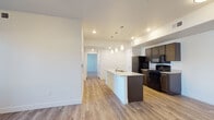 This well-appointed single-floor unit offers a thoughtfully designed open-concept layout that seamlessly connects the kitchen and living areas. The kitchen is a standout feature, centered around a sleek island with a built-in sink and light granite countertops, complemented by rich dark wood cabinetry and a full suite of black appliances including a refrigerator, electric range, and over-the-range microwave. Light wood-look plank flooring runs throughout the main living areas, enhancing the bright, clean aesthetic created by crisp white walls and recessed lighting with elegant pendant fixtures above the island. The living area benefits from natural light streaming through glass patio doors that open to a balcony, with track lighting adding additional illumination. The bedrooms are carpeted for comfort and each features a window allowing natural light to fill the space. Two bathrooms serve the unit, both finished with dark wood vanities, granite-style countertops, and light wood-look flooring. One bathroom includes a shower-tub combination, while the other offers a walk-in shower configuration. A hallway connects the bedrooms and bathrooms, providing a practical and well-organized flow throughout the unit.