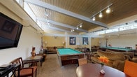 Located at 5825 Reseda Blvd in the Tarzana neighborhood of Los Angeles, this single-floor community clubhouse is designed to serve as a versatile social and recreational hub. The space is defined by its striking vaulted wood-plank ceilings with exposed beams, illuminated throughout by track lighting that creates a warm, inviting atmosphere.

The main gathering area is generously proportioned and anchors the space with a full-size pool table at its center, surrounded by comfortable lounge seating and framed artwork along the walls. A large flat-screen television mounted on the wall, paired with media storage and additional seating, provides a dedicated entertainment zone. High clerestory windows along the upper walls allow natural light to filter in, enhancing the openness of the room.

An adjacent dining and bar area features a wraparound counter with bar-height seating and round tables with colorful accent chairs, making it well-suited for casual gatherings and social events. Fresh floral arrangements add a welcoming touch throughout.

Near the entrance, a cozy lounge nook with a built-in fireplace and rich red upholstered seating offers a relaxed retreat, with glass doors providing views to the outdoor area beyond. The consistent use of warm tones, patterned carpeting, and thoughtful furnishings throughout creates a cohesive and comfortable communal environment.