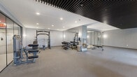 Located at 269 Orange Street in New Haven, CT, this fitness center offers a spacious, single-floor layout designed to support a comprehensive workout experience. The space is divided into distinct functional zones, each thoughtfully appointed with professional-grade equipment.

The strength training area features a power rack with weight plates, a cable machine, adjustable benches, and a full dumbbell rack, all set on durable carpet flooring. Full-length framed mirrors line the walls, enhancing the sense of space and allowing for proper form checks during lifts. Recessed lighting and a striking black slatted ceiling treatment add a sleek, modern aesthetic to this zone.

The cardio area is positioned along a windowed wall, providing natural light and views of the surrounding neighborhood. Multiple treadmills, stationary bikes, and additional strength machines are arranged to maximize the open floor plan. The black slatted ceiling feature continues here, creating a cohesive design language throughout.

A separate flex studio is enclosed by floor-to-ceiling glass partitions with black framing, offering a dedicated space for yoga, stretching, or mat-based workouts. The studio features light wood-tone flooring, textured gray accent walls, and exercise mats. A water station and waste receptacle are conveniently placed within the main floor. The overall ambiance is clean, contemporary, and functional.