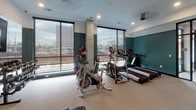 Located at 2512 Riverside Ave in Minneapolis, MN, this well-appointed fitness center offers a comprehensive workout environment on a single level. The space is designed with a modern aesthetic, featuring bold teal accent walls contrasted against crisp white ceilings and clean architectural lines. Recessed lighting throughout ensures a bright, energizing atmosphere, complemented by expansive floor-to-ceiling windows that flood the cardio area with natural light and provide open views of the surrounding cityscape.

The fitness floor is outfitted with a variety of cardio equipment, including treadmills, stationary bikes, and a stair climber, all arranged to take advantage of the natural light from the large windows. A dedicated strength and flexibility zone houses a rack of dumbbells, a weight bench, and stability balls in multiple sizes. Wall-mounted water fountains provide convenient hydration stations, and a large flat-screen television is mounted prominently for entertainment during workouts. Full-length mirrored walls along one side of the room enhance the sense of space while allowing users to monitor their form. Durable, cushioned flooring throughout provides comfort and safety. A ceiling fan adds to the comfort of the space, making this fitness center a functional and inviting facility.