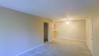 This single-floor residence offers a straightforward and functional layout designed for comfortable everyday living. The main living area is finished with wall-to-wall carpeting in a neutral tone, providing a warm and soft underfoot feel throughout. Smooth white ceilings are fitted with a ceiling fan, offering both ambient lighting and year-round climate comfort. The walls are painted in a soft beige hue, lending the space a calm and inviting atmosphere. A notable feature is the open pass-through counter that connects the living area to the kitchen, promoting an open feel and ease of interaction between spaces. The kitchen area, visible from the living room, includes upper cabinetry and a countertop overhang suitable for casual seating or serving. A walk-in closet is conveniently accessible from the main living space, offering generous built-in storage with an interior rod and shelving system. White baseboards line the perimeter of the rooms, adding a clean finishing detail. Overall, the space presents a simple, move-in-ready environment with practical features and a cohesive, neutral color palette throughout.