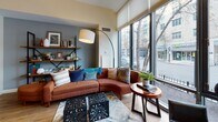 Located at 75 Station, this thoughtfully designed single-floor unit showcases a modern, open-concept layout that maximizes both space and functionality. The living area is anchored by expansive floor-to-ceiling windows that flood the interior with natural light and offer street-level views of the surrounding neighborhood, creating a bright and inviting atmosphere throughout.

The kitchen is efficiently designed with white cabinetry featuring glass-front upper doors, stainless steel appliances including a range, microwave, dishwasher, and refrigerator, along with light stone countertops. Warm yellow accent walls and track lighting add a vibrant touch to this culinary space.

A distinctive row of slim black vertical posts with frosted glass panels serves as an elegant divider between the sleeping area and the living room, providing a sense of separation while preserving the open, airy feel. The sleeping area features warm hardwood flooring, a built-in closet, and direct access to the hallway and entry.

The full bathroom is clean and contemporary, outfitted with a combination tub and shower, a white vanity with stone countertop, a large framed mirror, and neutral tile flooring. Throughout the unit, light hardwood floors, high ceilings, and a cohesive modern aesthetic create a comfortable and stylish living environment.