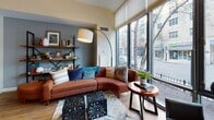 Located at 75 Station, this thoughtfully designed single-floor unit showcases a modern, open-concept layout that maximizes both space and functionality. The living area is anchored by expansive floor-to-ceiling windows that flood the interior with natural light and offer street-level views of the surrounding neighborhood, creating a bright and inviting atmosphere throughout.

The kitchen is efficiently designed with white cabinetry featuring glass-front upper doors, stainless steel appliances including a range, microwave, dishwasher, and refrigerator, along with light stone countertops. Warm yellow accent walls and track lighting add a vibrant touch to this culinary space.

A distinctive row of slim black vertical posts with frosted glass panels serves as an elegant divider between the sleeping area and the living room, providing a sense of separation while preserving the open, airy feel. The sleeping area features warm hardwood flooring, a built-in closet, and direct access to the hallway and entry.

The full bathroom is clean and contemporary, outfitted with a combination tub and shower, a white vanity with stone countertop, a large framed mirror, and neutral tile flooring. Throughout the unit, light hardwood floors, high ceilings, and a cohesive modern aesthetic create a comfortable and stylish living environment.