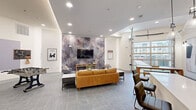 Located in Garland, TX, this single-floor community lounge and kitchen space offers a thoughtfully designed open-plan layout that seamlessly blends social and functional areas. The living room is anchored by a dramatic full-wall mural with an abstract stone-like pattern, serving as a bold backdrop for a wall-mounted flat-screen TV. Comfortable seating arrangements, including a tan leather sofa, gray accent chairs, and marble-topped coffee tables, create an inviting gathering space. A foosball table and shuffleboard add recreational appeal, making the area well-suited for social events. The kitchen is appointed with gray shaker-style cabinetry, white quartz countertops, and a textured brick-tile backsplash that adds warmth and character. Floating wood shelves provide open storage, while a stainless steel refrigerator and undermount sink complete the functional setup. A central island with bar seating serves as a natural gathering point. Glass pendant lights illuminate the workspace with style. Large garage-style windows flood the space with natural light and offer views of the surrounding exterior. Throughout, large-format gray tile flooring laid in a herringbone pattern ties the design together, contributing to the space's clean, contemporary aesthetic.