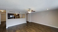 This single-story space in Fort Worth offers an open-concept layout that connects the living area to the kitchen, creating a functional and cohesive flow throughout. Dark hardwood flooring spans the main living area, providing a rich, grounded aesthetic that contrasts well with the neutral gray walls and crisp white baseboards. A ceiling fan with integrated lighting is centrally positioned overhead, contributing to both comfort and illumination in the space.

The textured popcorn ceiling adds a classic touch, while HVAC vents are thoughtfully integrated into the ceiling for efficient climate control. The kitchen is partially separated by a raised bar-height counter finished with granite-style countertops, offering a natural gathering point and casual dining option. Dark cabinetry lines the kitchen walls, complemented by stainless steel appliances, including a refrigerator and microwave, adding a modern contrast to the warm tones of the space.

Visible toward the back of the living area is a built-in shelving unit, providing practical storage or display space. The open passage nearby suggests additional rooms beyond, contributing to the overall sense of depth and utility within this well-appointed single-floor layout.