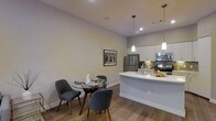 This single-floor residence showcases a thoughtfully designed open-concept layout that seamlessly connects the kitchen and dining areas, creating a bright and functional living environment. The kitchen is outfitted with sleek white cabinetry, stainless steel appliances including a refrigerator, microwave, and range, and a curved peninsula countertop that doubles as a prep and serving space. Pendant lights hang above the peninsula, adding a stylish focal point while providing targeted illumination. The dining area comfortably accommodates a round table with modern upholstered chairs, making it an inviting space for casual meals or entertaining. Warm hardwood-style flooring runs throughout, lending a cohesive and contemporary feel to the space. Neutral light grey walls provide a clean, versatile backdrop that complements the modern furnishings. Recessed lighting in the ceiling ensures consistent, well-distributed light throughout. A framed piece of wall art adds a personal touch to the dining nook. The overall ambiance is modern, clean, and welcoming, with a design that prioritizes both style and practicality in everyday living.