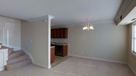 This two-story home showcases a thoughtfully designed open-concept layout that connects the main living, dining, and kitchen areas into one cohesive space. The living and dining area is finished with neutral-toned carpeting and painted in soft, warm beige tones, creating a welcoming and versatile atmosphere. Crown molding runs along the ceiling perimeter, adding a refined architectural touch throughout the space.

A decorative white column serves as a partial divider between the living area and the kitchen, providing subtle separation while maintaining an open feel. The kitchen features rich, dark cherry wood cabinetry paired with a tile floor, offering a warm contrast to the lighter tones of the adjacent living spaces. A sink with a standard faucet and a built-in dishwasher are visible, reflecting a practical and functional layout.

An elegant chandelier with a classic candelabra design anchors the dining area, serving as a standout lighting fixture. The staircase, partially visible at the edge of the space, provides access to the second floor. Overall, the space balances traditional design elements with an open, airy layout.