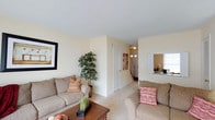 This two-floor residence in Charlestown offers a thoughtfully arranged layout designed for comfortable everyday living. The main level features a spacious living area finished in warm, neutral tones, with soft carpeting underfoot and light-colored walls that create a bright, inviting atmosphere. The living room accommodates generous seating arrangements and is accented by framed wall art and decorative greenery, adding character to the space. A wooden coffee table serves as a central anchor to the seating area, while a pass-through window connects to an adjacent room, enhancing the open feel of the main level. Natural light filters into the space, contributing to its airy ambiance. A staircase visible from the living area provides access to the upper floor, seamlessly integrating the two levels of the home. The entryway, glimpsed near the base of the stairs, features a transom window above the front door, allowing additional light into the foyer. Throughout the main level, the consistent use of neutral finishes and soft furnishings creates a cohesive, welcoming environment well-suited to a variety of lifestyles.