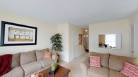 This two-floor residence in Charlestown offers a thoughtfully arranged layout designed for comfortable everyday living. The main level features a spacious living area finished in warm, neutral tones, with soft carpeting underfoot and light-colored walls that create a bright, inviting atmosphere. The living room accommodates generous seating arrangements and is accented by framed wall art and decorative greenery, adding character to the space. A wooden coffee table serves as a central anchor to the seating area, while a pass-through window connects to an adjacent room, enhancing the open feel of the main level. Natural light filters into the space, contributing to its airy ambiance. A staircase visible from the living area provides access to the upper floor, seamlessly integrating the two levels of the home. The entryway, glimpsed near the base of the stairs, features a transom window above the front door, allowing additional light into the foyer. Throughout the main level, the consistent use of neutral finishes and soft furnishings creates a cohesive, welcoming environment well-suited to a variety of lifestyles.