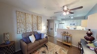 This single-floor residence offers a thoughtfully designed open-concept layout that connects the living area to the kitchen, creating a cohesive and functional living environment. The living room serves as the centerpiece of the space, featuring warm hardwood-style flooring, a ceiling fan with integrated lighting, and neutral gray walls that provide a versatile backdrop. A bold geometric gold wall art installation anchors the main wall, adding a striking decorative focal point that elevates the room's aesthetic. The living area is well-proportioned, offering comfortable space for both relaxing and entertaining.

The kitchen is accessible via a half-wall pass-through, giving the space an open and connected feel. It is outfitted with white cabinetry, a tile backsplash, stainless-style appliances including a microwave and refrigerator, and track lighting overhead. The half-wall also provides a convenient surface for casual dining or serving. A hallway visible from the living room leads to additional areas of the residence, hinting at a practical and well-organized floor plan. Overall, the space combines contemporary design elements with a warm, inviting ambiance suited for everyday living.