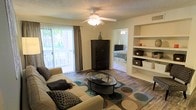 This single-floor space offers a well-organized and functional layout with a comfortable, casual ambiance. The living area serves as the heart of the home, featuring hardwood-style flooring that adds warmth and continuity throughout. A ceiling fan with a built-in light fixture provides both air circulation and ambient lighting, complementing the abundant natural light that streams in through a large sliding glass door dressed with vertical blinds and drape panels. The sliding door provides a view of outdoor greenery, enhancing the bright and airy feel of the room.

Built-in shelving units along one wall offer practical display and storage space, adding an architectural detail that contributes to the room's functionality. An open passageway connects the living area directly to the bedroom, which also features its own ceiling fan, creating a cohesive flow between spaces. The bedroom is partially visible and appears well-proportioned. The textured ceiling adds a classic residential touch throughout. Overall, the space presents a neat, livable environment with a relaxed, neutral aesthetic that is practical and inviting.