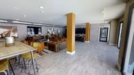 This single-floor communal space offers an open, airy layout with a contemporary industrial aesthetic that blends comfort and functionality. The flooring throughout is a light gray wood-look tile that lends a clean, modern feel to the expansive room. Structural columns wrapped in a warm golden-yellow finish serve as bold architectural accents, adding visual interest and helping to define distinct zones within the open plan.

The lounge area is anchored by a plush leather sofa and complemented by vibrant orange and yellow accent chairs arranged around a central coffee table atop a navy and white striped area rug. This seating zone is further enriched by a large decorative wall painting and a coordinating floor lamp, creating an inviting and stylish gathering spot.

Exposed brick walls along the perimeter reinforce the industrial character of the space, while recessed ceiling lighting provides consistent, even illumination throughout. Toward the rear, a dedicated dining or collaborative work zone features high-top seating and dark furnishings, offering a functional contrast to the relaxed lounge area. A corridor visible in the background suggests additional amenities or spaces beyond. Overall, the design strikes a balance between energetic, modern style and practical, multi-use functionality.