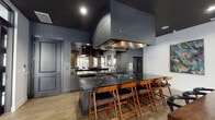 This sophisticated single-floor space showcases a seamlessly connected open-concept design that blends a fully equipped kitchen, a comfortable lounge area, and a formal dining zone into one cohesive environment. The kitchen is a standout feature, anchored by a dramatic oversized island with striking black marble countertops and lined with mid-century modern barstools in warm walnut tones. A large stainless steel range hood serves as an architectural focal point overhead, while dark cabinetry, mirrored backsplash accents, and a full suite of professional-grade appliances complete the culinary space. Adjacent to the kitchen, the dining area is centered around a substantial rustic wood communal table surrounded by sleek black chairs, with a trio of large abstract paintings displayed prominently on the white wall above. The lounge area features an inviting seating arrangement with leather and upholstered chairs, a tiered open shelving unit filled with greenery, and a wall-mounted television. Light wood-tone flooring flows throughout, providing a warm contrast to the deep charcoal ceilings and crisp white walls. Floor-to-ceiling glass partitions and large windows fill the space with natural light and offer views of an outdoor terrace, creating an airy, connected feel throughout.