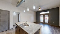 Located at 612 E Hickory St in Denton, TX, this modern 2-bedroom, 2-bathroom unit offers a well-designed single-floor layout with contemporary finishes throughout. The open-concept kitchen and living area serves as the heart of the space, showcasing soaring ceilings with exposed industrial ductwork, sleek pendant lighting, and wood-look flooring. The kitchen is equipped with granite countertops, a center island with a double sink, white upper cabinetry, contrasting gray lower cabinets, a tile backsplash, and stainless steel appliances including a refrigerator and built-in microwave. The living area features French doors that invite abundant natural light and provide outdoor views, complemented by a ceiling fan overhead and a bold accent wall. Both bedrooms are carpeted and include ceiling fans and windows with blinds that allow for natural light. The primary bedroom offers a generous footprint with dedicated closet space. The two bathrooms are finished with granite-topped dark wood vanities, wood-look tile flooring, and modern lighting fixtures. One bathroom features a full soaking tub with floor-to-ceiling gray tile surround. Three closets and a hallway complete the functional layout of this well-appointed Denton residence.