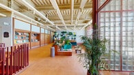 Located at 1525 N American St in Philadelphia, PA, the Loving Room is a thoughtfully designed community lounge spanning a generous single floor. The space showcases an industrial-modern aesthetic, highlighted by exposed concrete ceilings with visible structural beams and ductwork painted in crisp white, lending the interior an open, airy character.

Warm terracotta floor tiles extend throughout, providing a grounding contrast to the cool industrial ceiling above. A dramatic floor-to-ceiling curtain wall of large-pane windows floods the space with natural light and offers open views of the surrounding neighborhood streetscape. Beneath the windows, a continuous built-in teal bench seat lined with an abundant collection of potted plants creates a verdant, welcoming atmosphere.

The layout includes a distinct lounge area furnished with teal modular sofas and low wooden coffee tables, a curated bookshelf displaying reading materials, and wood-paneled built-in cabinetry along the walls. A glass-partitioned Work Lounge area is accessible via bold red-framed doors, offering a more enclosed setting for focused use.

A striking red-painted open staircase with metal railings leads to a leasing office on the lower level. A mural featuring botanical motifs adds an artistic accent near the stairwell. The overall ambiance balances industrial architecture with warm, natural materials and lush greenery, creating an inviting and functional communal environment.
