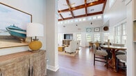 Located at 1701 Dawsonville Hwy in Gainesville, GA, this thoughtfully designed single-floor space makes a striking impression from the moment you enter. The combined kitchen and living room serves as the heart of the layout, anchored by a soaring vaulted ceiling with rich exposed wood beams and a coffered detail that draws the eye upward. A floor-to-ceiling stacked stone fireplace commands the living area, complemented by warm hardwood flooring and an abundance of natural light streaming through rows of large windows and clerestory transoms above. The kitchen features warm honey-toned wood cabinetry, granite-style countertops, a green subway tile backsplash, and a window above the sink that brings in views of the outdoors. The open-plan living area flows seamlessly into a sunlit lounge zone lined with multi-pane windows overlooking an outdoor deck, creating an airy, welcoming atmosphere perfect for gathering. A separate dining room offers an intimate setting with neutral tones, a round glass-top table, and woven window treatments that filter soft natural light. Throughout the space, consistent use of hardwood flooring, crisp white walls, and carefully curated furnishings create a cohesive, upscale aesthetic that balances warmth with elegance.