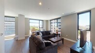 This single-floor urban residence is designed with a modern aesthetic, combining functionality with contemporary style throughout its well-organized layout. The open-concept living and dining area serves as the heart of the space, anchored by hardwood flooring and large floor-to-ceiling windows that flood the interior with natural light and frame expansive city views, including surrounding high-rise buildings and street-level activity below.

The living area comfortably accommodates a full seating arrangement centered around a rectangular coffee table, with a wall-mounted flat-screen television positioned on a sleek media console. Adjacent to the living area, a dedicated dining space features a dark wood table and chairs, positioned directly beside the oversized windows to maximize the urban panorama.

The kitchen is efficiently designed with dark cabinetry, light stone countertops, a white tile backsplash, and stainless steel appliances, including a range, over-the-range microwave, and dishwasher. A large window in the kitchen further enhances the sense of openness.

The bedroom is a calm, understated retreat with neutral tones, carpeted flooring, a dark wood bed frame with matching nightstands, and a window providing natural light. The bathroom features light tile flooring, a dark vanity with a stone countertop, and a clean, minimal design.