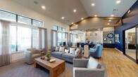 Located at 19770 Clay Rd in Katy, TX, this single-story community clubhouse showcases a thoughtfully designed open-concept interior that blends contemporary style with welcoming comfort. The main lounge area features soaring vaulted ceilings with recessed lighting, wide floor-to-ceiling windows dressed in sheer neutral drapery, and an abundance of natural light that flows throughout the space. Warm hardwood flooring extends across much of the interior, complemented by plush area rugs that define the seating zones. The lounge is furnished with generous gray sofas, accent chairs in soft blue tones, and a rustic wood coffee table, creating an inviting gathering environment. A bold navy blue accent wall adorned with curated artwork adds a striking focal point, while a wooden credenza with decorative lamps anchors the space. Adjacent to the lounge is a dining and meeting area featuring a large central table with bar-height seating, dramatic geometric pendant lighting, and a textured white brick accent wall. Dark cabinetry and a countertop wet bar complete this functional zone. Large windows throughout offer direct views of the outdoor pool and courtyard area, seamlessly connecting the interior to the surrounding community amenities. The overall ambiance is modern, polished, and community-focused.