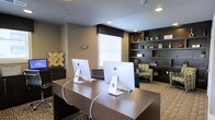 Located at 8110 Six Forks Rd in Raleigh, NC, this single-floor business center offers a professionally designed workspace tailored for productivity and comfort. The space features a sleek, contemporary aesthetic with a neutral color palette of warm taupes and deep charcoal tones that create a focused and polished atmosphere.

The main workstation area is anchored by a large, dark wood desk equipped with multiple iMac computers, providing residents or visitors with dedicated computing resources. Recessed lighting illuminates the space evenly from above, while a window with blinds allows for natural light and ventilation control.

Along the back wall, a built-in dark shelving unit spans floor to ceiling, offering ample display and storage space for books, décor, and personal items. Adjacent to the shelving, a cozy lounge chair with patterned upholstery provides a relaxed reading or waiting area, adding a residential warmth to the otherwise professional setting.

Decorative wall art and accent pieces contribute to the space's curated character, while the textured carpeting underfoot adds both comfort and visual interest. The thoughtful layout balances functionality with design, making this business center a practical and inviting destination for work or study.