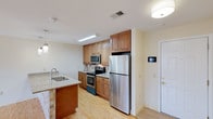 This single-floor space showcases a thoughtfully designed kitchen that serves as its centerpiece. The kitchen is outfitted with a full suite of stainless steel appliances, including a French-door refrigerator, an electric range with oven, and an over-the-range microwave, all complemented by a classic tile backsplash that adds texture and visual interest. Warm-toned wood cabinetry extends from floor to ceiling, providing generous storage space, while light granite countertops offer a bright contrast. A kitchen island with a matching granite surface houses a double undermount sink, creating a functional and social focal point in the space. Light hardwood flooring runs throughout, contributing to a clean, cohesive aesthetic. Recessed and flush-mount ceiling lighting ensures the space is well-lit. Near the entrance, a white paneled door and a wall-mounted control panel suggest smart or climate-control integration. The overall ambiance is bright, functional, and modern, with warm wood tones and light-colored walls working together to create an inviting and practical environment.