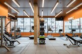 This fitness center is a thoughtfully designed, single-floor facility that balances functionality with striking visual appeal. The space is defined by bold orange accent walls and a dramatic floor-to-ceiling abstract mural that serves as a dynamic focal point, creating an energizing atmosphere throughout. Linear LED lighting installed across a dark, wood-paneled ceiling provides bright, uniform illumination across the entire floor.

Large floor-to-ceiling windows line the street-facing wall, flooding the interior with natural light and offering open views of the surrounding urban streetscape. The flooring consists of durable, large-format rubber tiles suited for high-impact activity.

The cardio zone features a row of treadmills, an elliptical, a rowing machine, and a stationary bike, all positioned to take advantage of the natural light from the windows. A dedicated strength training area includes a multi-station cable machine, an adjustable weight bench, and a full dumbbell rack. Functional training accessories are neatly organized on a freestanding storage rack, including medicine balls, stability balls, kettlebells, and exercise mats.

Full-length mirrors are strategically placed to enhance the sense of space and support proper form during training. A concrete structural column anchors the center of the room, adding an industrial character to the overall design. The result is a comprehensive, well-organized fitness facility with a modern and motivating aesthetic.