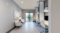 This contemporary single-floor space is thoughtfully designed to serve as a professional business center, seamlessly blending productivity with comfort. The layout is divided into two distinct functional zones that work harmoniously together.

On one side, a row of built-in workstations lines the wall, each framed by elegant wood-trimmed dividers with a textured stone-like finish. Ergonomic white task chairs accompany each station, and stylish pendant lights hang overhead to provide focused illumination. A large monitor is mounted at the end of the workspace, making the area well-equipped for a variety of professional tasks.

The opposite side of the room features a relaxed lounge area with a pair of curved, cream-upholstered accent chairs and a sleek floor lamp, ideal for informal meetings or quiet reading. A large abstract artwork adorns the soft grey wall, adding a refined artistic touch to the space.

Large black-framed glass doors at the far end of the room flood the interior with natural light while offering a view of lush greenery outside. Light grey tile flooring runs throughout, tying the two zones together cohesively. Recessed ceiling lighting complements the natural light to ensure the space remains bright and inviting throughout the day.