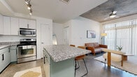 Located at 888 N Fourth St in Phoenix, AZ, this single-floor unit offers three bedrooms and two bathrooms within a thoughtfully designed, compact layout. The open-concept kitchen and living area serve as the heart of the space, anchored by a sage green island with granite countertops and bar seating for two. The kitchen is fully equipped with stainless steel appliances, including a refrigerator, dishwasher, microwave, and range, complemented by white upper cabinetry and sage lower cabinets. Polished concrete floors extend throughout the main living area, adding an industrial character that is echoed by exposed concrete ceiling elements visible in the living room and bedrooms. The living room benefits from natural light through large vertical-blind windows and offers a comfortable, open feel. Each bedroom features plush carpet flooring, closet space, and windows that bring in natural light. The bathrooms are finished with granite-topped vanities and white tile surrounds. The overall aesthetic blends modern urban design with warm, functional finishes, creating a cohesive and practical living environment in the heart of Phoenix.