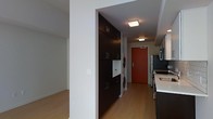 This single-floor space showcases a contemporary design with a cohesive, modern aesthetic throughout. The kitchen is equipped with stainless steel appliances, including a refrigerator and microwave, and features dark cabinetry paired with light countertops, creating a striking contrast that adds visual depth. The layout flows naturally into an entryway corridor, where a bold red front door serves as a distinctive focal point, lending a pop of color to the otherwise neutral palette.

A standout feature of this space is the in-unit laundry area, which houses a stacked washer and dryer neatly integrated alongside floor-to-ceiling dark cabinetry. This built-in storage solution maximizes functionality while maintaining a clean, organized appearance. The cabinetry features sleek hardware and extends to ceiling height, offering generous storage capacity.

Warm hardwood-style flooring runs continuously throughout the space, contributing to a cohesive and inviting atmosphere. Recessed and flush-mounted ceiling lights provide practical illumination across all areas. The open layout between the living area and the kitchen corridor enhances the sense of flow and spaciousness. Overall, the space combines practical amenities with a polished, urban aesthetic suited for modern living.