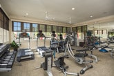 This fitness center in Columbus, Georgia offers a comprehensive workout environment thoughtfully designed for a full range of exercise activities. The single-floor space is finished with neutral carpeting and warm beige walls accented by crown molding, giving it a polished, welcoming atmosphere. Large multi-pane windows line the perimeter, allowing abundant natural light to fill the room while providing views of the surrounding outdoor greenery. Roller shades offer adjustable light control throughout the day.

Full-length wall mirrors are strategically positioned to visually expand the space and allow users to monitor their form during exercise. Two ceiling fans help maintain comfortable airflow. Recessed lighting supplements natural light for consistent illumination.

The equipment selection is extensive, including treadmills, an elliptical trainer, and a stationary bike for cardiovascular workouts. Strength training is well supported with a Cybex cable machine, a multi-function weight station, adjustable benches, and a full dumbbell rack. Functional training tools such as resistance bands, kettlebells, medicine balls, and stability balls round out the offerings. A French door provides direct access to the outdoors, and posted fitness rules and instructional signage contribute to an organized, professional environment.
