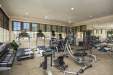 This fitness center in Columbus, Georgia offers a comprehensive workout environment thoughtfully designed for a full range of exercise activities. The single-floor space is finished with neutral carpeting and warm beige walls accented by crown molding, giving it a polished, welcoming atmosphere. Large multi-pane windows line the perimeter, allowing abundant natural light to fill the room while providing views of the surrounding outdoor greenery. Roller shades offer adjustable light control throughout the day.

Full-length wall mirrors are strategically positioned to visually expand the space and allow users to monitor their form during exercise. Two ceiling fans help maintain comfortable airflow. Recessed lighting supplements natural light for consistent illumination.

The equipment selection is extensive, including treadmills, an elliptical trainer, and a stationary bike for cardiovascular workouts. Strength training is well supported with a Cybex cable machine, a multi-function weight station, adjustable benches, and a full dumbbell rack. Functional training tools such as resistance bands, kettlebells, medicine balls, and stability balls round out the offerings. A French door provides direct access to the outdoors, and posted fitness rules and instructional signage contribute to an organized, professional environment.